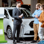 Sales manager showing car charging station to a young couple, selling electric cars in the showroom. Concept of buying eco-friendly car for family