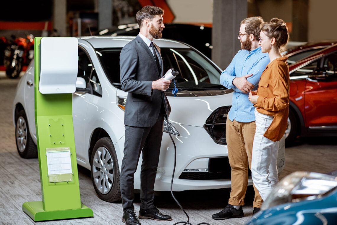 Sales manager showing car charging station to a young couple, selling electric cars in the showroom. Concept of buying eco-friendly car for family